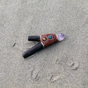 Peut inclure: Une pipe en bois avec un enroulement en cuir et trois accents de pierres précieuses. La pipe est décorée d'un cristal d'améthyste violet à l'extrémité.