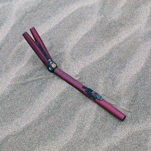 Könnte beinhalten: Ein dunkelroter Holzstab mit schwarzen und goldenen Dekor-Elementen und einem türkisfarbenen Edelstein. Der Stab liegt auf einer sandigen Oberfläche.