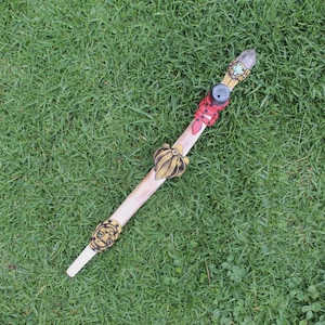May include: A wooden wand with a clear crystal tip, adorned with gold and red accents. The wand has a decorative design with a face and a leaf motif.