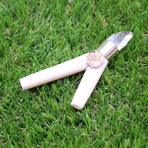 May include: A cream-coloured, two-piece tool with a silver band and a clear crystal at one end. The tool has a circular, patterned design in the centre. The tool is resting on a bed of green grass.