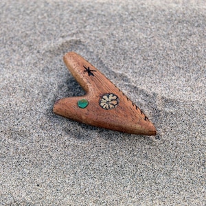 Könnte beinhalten: Ein herzförmiger Anhänger aus Holz mit einem grünen Stein und einem geschnitzten Blumendesign. Der Anhänger liegt auf einer sandigen Oberfläche.