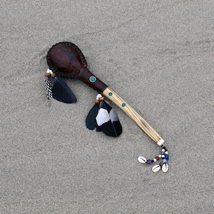 May include: A brown leather and wood rattle with black and white feathers and beads. The rattle is lying on a sandy surface.