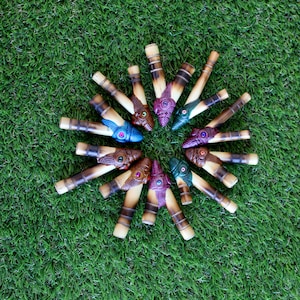 May include: A set of 12 wooden blowguns with colorful painted designs. Each blowgun has a different color and design, and they are arranged in a circle on a green surface.