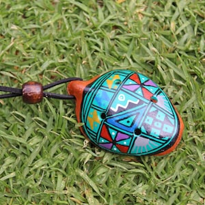 Op de afbeelding: Een kleurrijke keramische ocarina-hanger met een geometrisch ontwerp in tinten blauw, groen, rood, geel en paars. De ocarina is geregen aan een zwart koord met een bruine houten kraal.