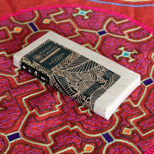 May include: A brown paper wrapped bar of dark chocolate with a black and gold label. The label reads "Kawsay Cacao Ceremonial Cacao From the Amazon Rainforest Pucallpa Ucayali Peru 200g Peso Neto".