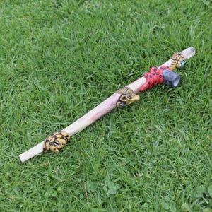 May include: A decorative wooden pipe with animal-themed carvings and embellishments. The pipe features a light-colored wooden body with gold and red accents, and a dark gray bowl. The carvings include a snake, a bird, and a ram.