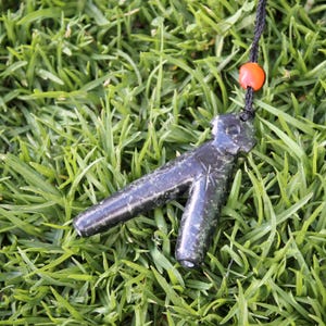 May include: A dark green stone pendant shaped like a stylized human figure with a star carved into the chest. The pendant is hanging from a black cord with an orange bead.
