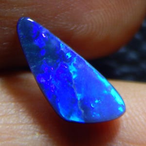 May include: A vibrant blue opal gemstone with a triangular shape. The stone displays a mesmerizing play of color, with flashes of bright blue and hints of other hues. The opal is held between fingers, showcasing its natural beauty.