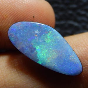May include: A polished, oval-shaped opal gemstone. The opal displays a vibrant play-of-color, with flashes of blue, green, and red hues across a predominantly blue background. The stone is held between fingers, showcasing its natural beauty.