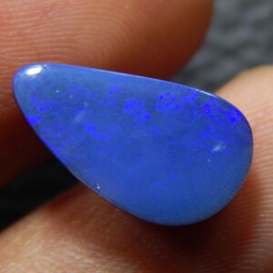 May include: A polished, teardrop-shaped opal gemstone. The stone has a deep blue color with flashes of vibrant blue and purple. The opal is held between fingers, showcasing its natural beauty and unique color play.