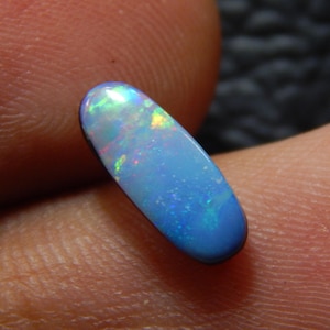 May include: A polished, oval-shaped opal gemstone. The opal displays a vibrant play-of-color, with flashes of blue, green, and yellow against a pale blue base. The gemstone is held between fingers, showcasing its iridescent qualities.