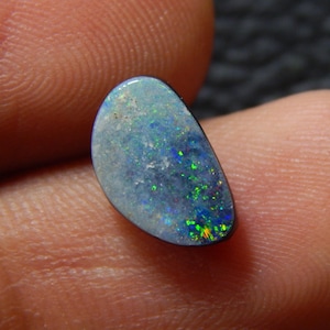 May include: A polished, oval-shaped opal gemstone. The opal displays a vibrant play-of-color, with flashes of green, blue, and yellow set against a base of light blue and black. The gemstone is held between fingers.