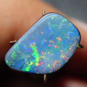 May include: A gold ring featuring a large, iridescent opal. The opal has a blue base color with flashes of green, red, and yellow. The stone is irregularly shaped and set in a simple gold band.