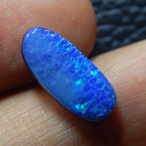 May include: A vibrant blue opal gemstone with a polished, oval shape. The stone displays a mesmerizing play of color, with flashes of bright blue and hints of green. The opal is held between two fingers, showcasing its natural beauty.