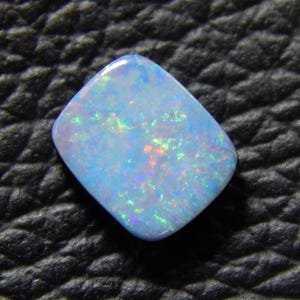 May include: A polished, blue opal gemstone with a rounded, square shape. The opal displays a play of color with flashes of green, orange, and purple. The gemstone is set against a textured black surface.