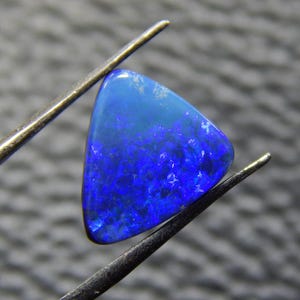 May include: A triangular, polished gemstone with a vibrant blue color gradient. The gem is held by metal tweezers, showcasing its iridescent surface. The stone's color transitions from a lighter blue at the top to a deep, rich blue at the base.