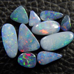 May include: A collection of polished opal gemstones in various shapes, including oval, teardrop, and triangular forms. The opals display a play of color with iridescent flashes of blue, green, red, and yellow. The gemstones are set against a dark, textured background.