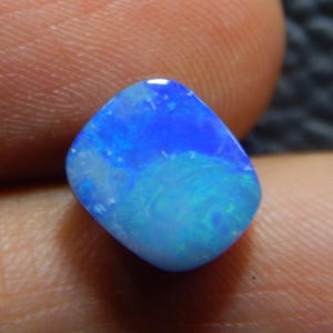 Può includere: Una gemma di opale lucidata, che mostra una gamma di blu e verdi. La superficie della pietra riflette la luce, creando un motivo a vortice di colore. La gemma è tenuta in una mano, mostrando le sue dimensioni e la sua forma.