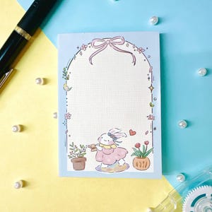 May include: A blue notepad with a white bunny watering plants in a garden setting. The bunny is wearing a pink dress and a white headband. The notepad has a white border with a pink bow at the top.