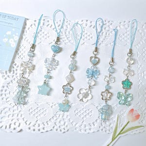 May include: Six phone charms featuring light blue and clear beads, bows, stars, hearts, and butterflies on thin blue cords. The charms are arranged vertically, showcasing various bead shapes and metallic accents in a cohesive light blue color palette.
