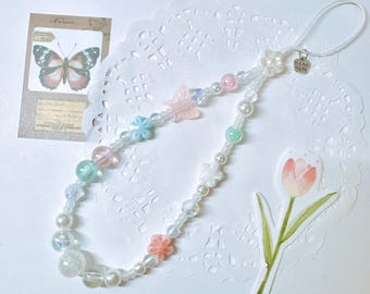 May include: A white phone charm with a beaded chain featuring pastel colored flowers, beads, and a small butterfly charm. The charm also includes a small silver tag with the text "Hand Made".
