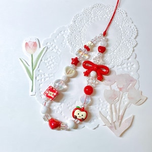 May include: A beaded phone charm featuring red, white, and clear beads, with red heart, star, and bow accents. Includes a smiling apple charm and a red cord for attachment. The charm is on a white lace doily.