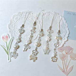 May include: Five white beaded phone charms with various charms, including hearts, bows, and flowers. Each charm has a silver-colored clasp and a white cord. The charms are arranged on a white doily with floral illustrations.
