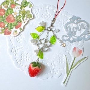 May include: A charm bracelet with a silver clover charm, green leaves, white flowers, and a red strawberry charm. The bracelet is attached to a red string.