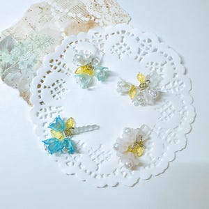 May include: Four pairs of delicate flower-shaped earrings made of clear, white, and blue resin. The earrings are arranged on a white doily.