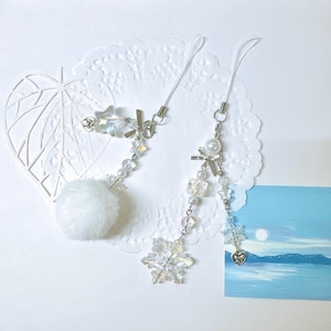 May include: Two white and silver charm phone straps with snowflake and pom-pom accents. The charms are hanging from a white background.