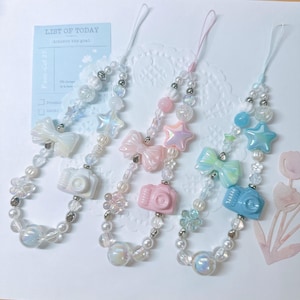 May include: Three pastel-colored phone charms with pearl beads, bows, stars, and camera charms. The charms are on a white background.