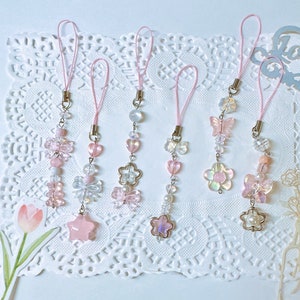 May include: Five pink and white beaded phone charms with different designs, including butterflies, flowers, and stars. Each charm is attached to a pink string.