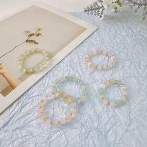 May include: Five pastel-colored beaded bracelets with gold accents. The bracelets are arranged on a light blue textured surface.