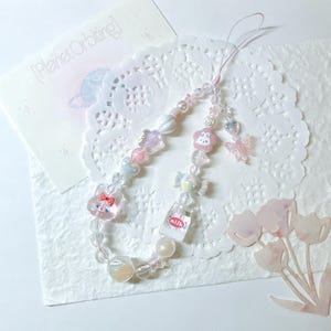 May include: A beaded phone charm with a variety of pastel-colored beads and charms. The charm includes a cat, flower, butterfly, and milk carton charm. The background includes a card with the text "Planet Orbiting".