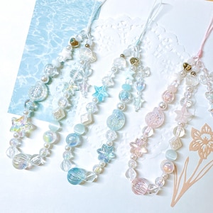 May include: Three colorful beaded phone charms with iridescent and pastel beads, including stars, hearts, and shells. The charms are attached to white cords.