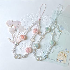 May include: Three beaded phone charms with clear, iridescent, and pastel-colored beads. Each charm features a loop for attachment and decorative elements like stars. The charms are arranged on a white, lace-like surface.