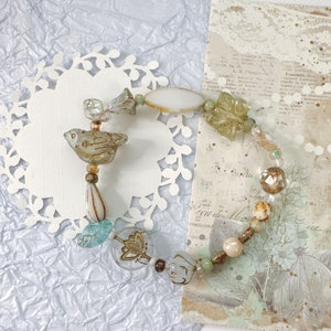 May include: A delicate bracelet made of glass beads in shades of white, blue, and green. The bracelet features a small bird charm and a floral charm. The bracelet is displayed on a white doily.