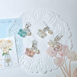 May include: Four pairs of clear earrings with floral and butterfly designs. The earrings feature various colors, including blue, pink, and clear, with pearl accents. The earrings are displayed on a white doily with floral illustrations.