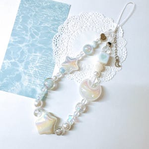 May include: A white and blue phone charm with a crescent moon, stars, and beads. The charm is attached to a white string.