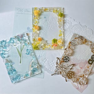May include: Three rectangular resin photo frames. One frame features yellow and green floral accents with pearls. Another frame contains blue crystals and small white shells. The third frame displays silver-toned metal gears and a butterfly charm.