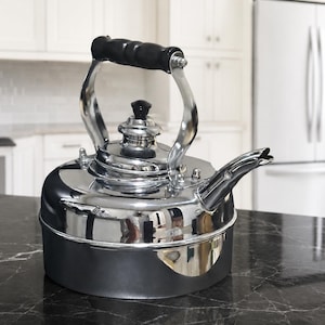 May include: A stovetop kettle with a polished chrome finish on the upper body and a matte black base. It features a curved spout, a removable lid with a black knob, and a black handle attached to the chrome frame. The design is rounded and metallic.