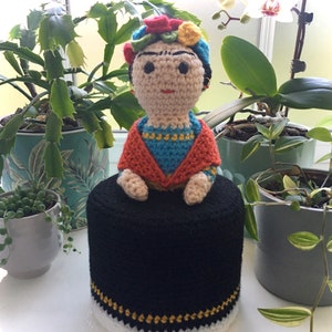 May include: Crocheted doll with a black, white, and gold crocheted hat. The doll is wearing a red, blue, and yellow crocheted outfit. The doll is sitting on a black crocheted base.