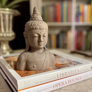 May include: A gray stone bust of Buddha sits on top of two books. The top book is titled "The Most Beautiful Libraries" and the bottom book is titled "The Most Beautiful Opera Houses".