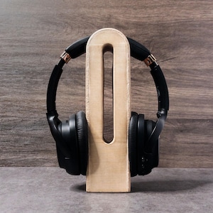 May include: Black over-ear headphones with rose gold accents rest on a light-colored wooden headphone stand. The stand has a rectangular base and a tall, open-center design. The background is a wooden wall.