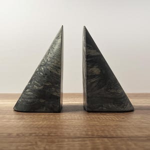 May include: A pair of triangular bookends with a marbled grey and black pattern. The bookends are made of a solid material and are set on a wooden surface. They are designed to hold books upright.
