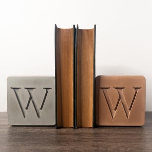 May include: Two concrete bookends shaped like the letter "W" are holding up two books with brown spines. The bookends are gray and brown.