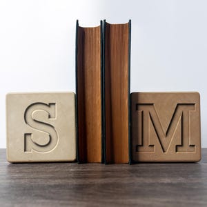 May include: Two hardback books stand between two square bookends. One bookend is beige with a carved "S", the other is brown with a carved "M". The books have brown pages and dark green spines.