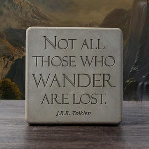 May include: A square, gray stone block with the quote "Not all those who wander are lost" by J.R.R. Tolkien, carved into the surface. The block is set against a backdrop of a mountain landscape.