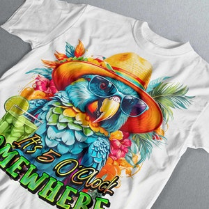 May include: White t-shirt featuring a colourful parrot wearing a straw hat and sunglasses. The parrot is surrounded by tropical flowers and leaves. The text "It's 5 O'clock Somewhere" is printed on the shirt.