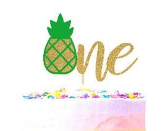May include: A frosted cake with colorful sprinkles on a pink cake stand. A green and gold pineapple cake topper and gold glitter text that reads "one" are on top of the cake. The cake has a gradient of colors.